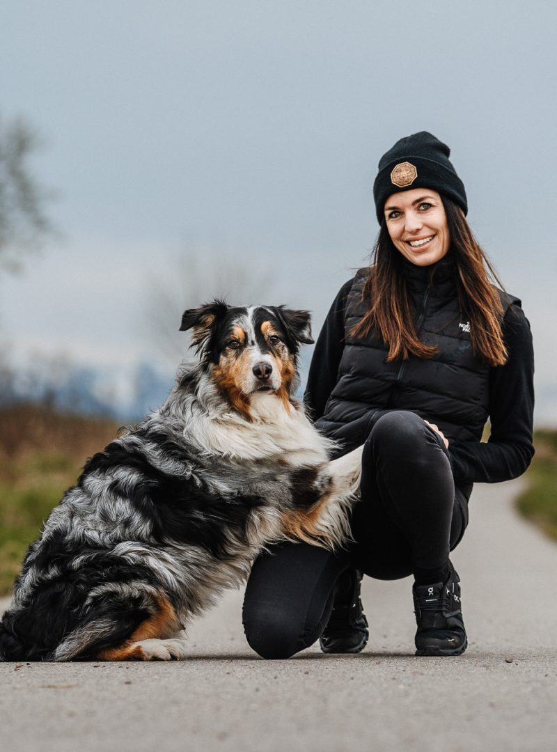 Trainerin kniet auf einem Feldweg neben ihrem Australian Shepherd